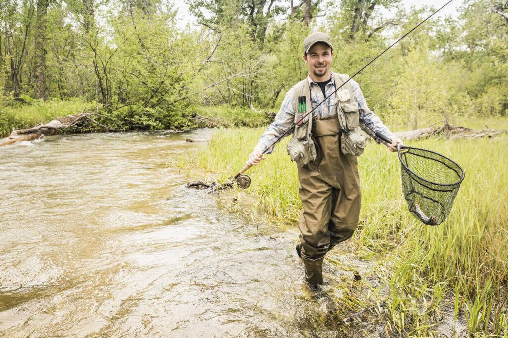 Man in river wearing best waders carrying fishing net .